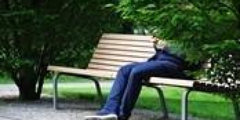 Ağaçların ve çimlerin olduğu bir parkta, yol kenarında bir bankta gövdesinin üst tarafı ağaçtan görünmeyen bir kişi oturuyor. Lacivert kot pantolon, lacivert mont ve mavi spor ayakkabıları var. Elindeki telefonla meşgul.