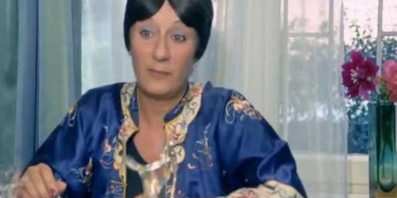 Sevda Aydan mavi ve desenli bir sabahlık giymiş, masada oturuyor. Kısa siyah saçları var ve yüzü ciddi bir ifade taşıyor. Masanın üzerinde bir bardak ve başka cam eşyalar bulunuyor. Arka planda, pencere önünde vazoda çiçekler var. Pencerenin dışında bulanık bir manzara görünüyor.