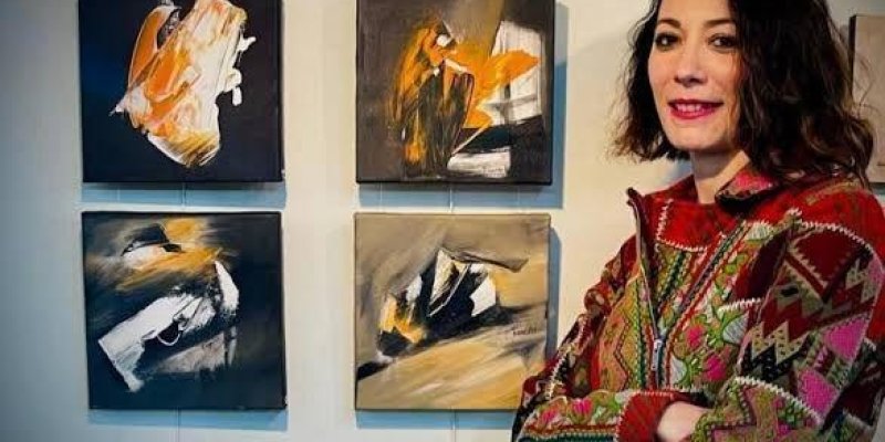 Bir sanat galerisi veya sergi alanı. Kısa saçlı Özge Gökbulut Özdemir, kollarını kavuşturmuş, üzerinde renkli desenli bir ceket var. Arka planda duvarda asılı dört adet küçük kare tablo bulunuyor. Bu tablolar soyut tarzda ve ağırlıklı olarak siyah, beyaz, turuncu ve gri tonlarında. Tabloların önünde durarak kameraya gülümsüyor.