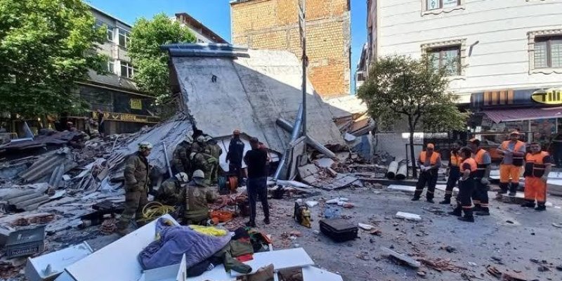 Yıkılmış bina enkazı ve çevresinde çalışan ekipler. Ön planda yere yığılmış beton, tuğla ve çeşitli moloz parçaları, aralarında dağınık şekilde alet çantaları, hortumlar ve örtülere sarılı malzemeler. Solda koruyucu kıyafetli birkaç itfaiye ya da arama‑kurtarma görevlisi enkazın önünde toplanmış Ortada enkazın üzerine doğru eğilmiş büyük, tek parça halinde devrilmiş bir beton duvar, etrafında bükülmüş metal direkler ve kırık yapı elemanları.