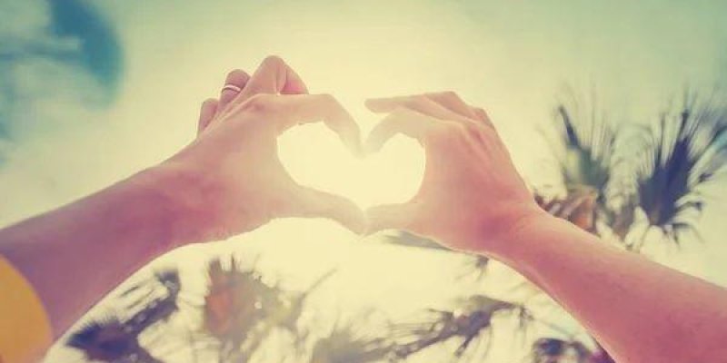 Gökyüzüne doğru uzatılmış iki elin parmaklarıyla kalp şekli yaptığı bir fotoğraf. Kalp şeklinin ortasında parlak güneş ışığı var ve ışık parmakların arasından yayılıyor. Eller açık tenli; sol elde bir yüzük. Arka planda mavi gökyüzü, hafif bulutlar ve alt kısımda bulanık palmiye ağaçları siluetleri.