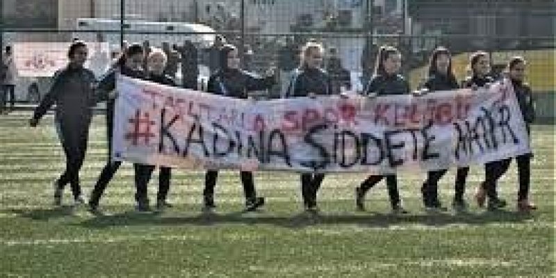 Çim sahada 7-8 genç kadın, siyah yazılı beyaz “kadına şiddete hayır” pankartı açmışlar.