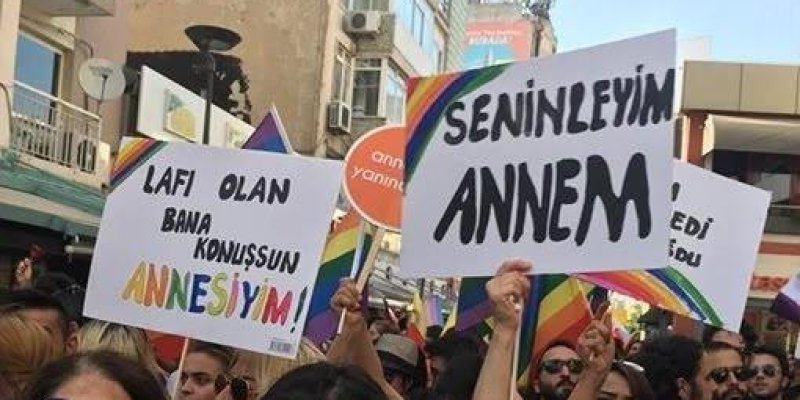Kalabalık bir yürüyüşte gökkuşağı bayrakları taşıyan insanlar. Ön planda iki beyaz pankart tutuluyor. Soldaki pankartta siyah ve renkli harflerle “Lafı olan bana konuşsun ANNesiyim!” yazısı. Sağdaki pankartta büyük siyah harflerle: “SENİNLEYİM ANNEM” yazıyor.