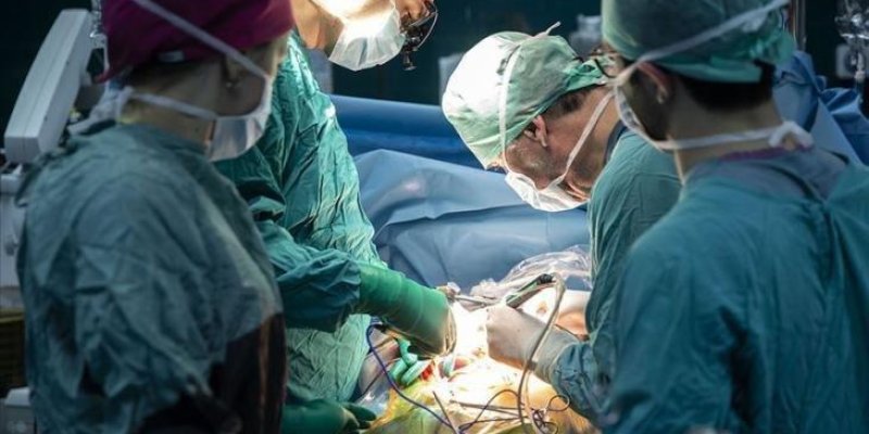 Ameliyat masasında yatan bir hasta etrafında toplanmış cerrahi ekip. Yeşil steril cerrahi kıyafetler ve bone takmış, maske ile yüzleri kapalı en az dört sağlık çalışanı operasyon yapıyor. Orta kısımda iki cerrah, parlak ameliyat lambasının ışığı altında hastanın göğüs-belin bulunduğu bölge üzerinde çalışıyor; ellerinde cerrahi aletler var. Ön plandaki hasta, mavi cerrahi örtülerle büyük ölçüde kaplı; yalnızca ameliyat edilen bölge açıkta bırakılmış. Masada hastaya bağlı birkaç ince hortum ve kab