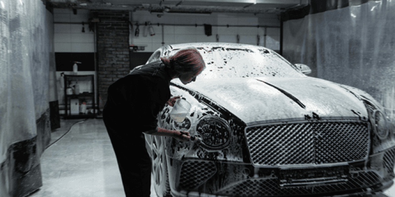 Oto yıkama istasyonunda çalışan bir kişi. Koyu renkli iş kıyafetleri giymiş ve pembe saçları var. Ellerinde bir sünger tutarak, bol köpüklü beyaz bir Bentley marka aracın ön kısmını yıkıyor. Arka plan gri tonlarda ve hafif loş bir ortamda çekilmiş. Aracın ön ızgarası ve büyük farları var. Yerler ıslak ve köpüklü.