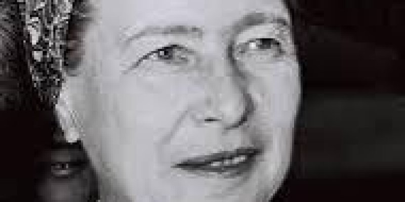 Ünlü Fransız yazar ve filozof Simone de Beauvoir’in, siyah beyaz bir portre fotoğrafı. Başına desenli bir eşarp bağlamış, yüzü hafifçe sağa dönük ve hafif bir gülümseme ile ileriye bakıyor. Üzerinde çizgili bir ceket var ve arka planda başka bir kişi bulanık bir şekilde görünüyor.