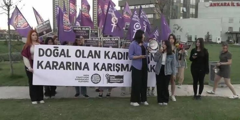 Bir grup insan, açık havada bir araya gelmiş ve pankartlar taşıyor. Pankartın üzerinde "DOĞAL OLAN KADINLARIN KARARINA KARIŞMAMAK" yazıyor. Pankartın altında "CİNAYETLERİ DURDURACAĞIZ PLATFORMU" ve "GENÇ FEMİNİSTLER" logoları bulunuyor. Arkada, mor renkli bayraklar taşıyan insanlar var, bayrakların üzerinde kadın sembolleri ve "DOĞAL OLAN NORMA DEMOKRASİ" yazıları yer alıyor. Katılımcılar arasında mikrofon ve megafon kullanan kişiler de var. Arka planda yü