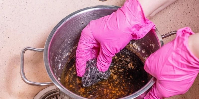 Mutfak lavabosunda, kirli sıvı ve yemek artıkları olan paslanmaz çelik tencerede koyu renkli, yağlı ve parçacıklı bir su karışımı; tencerenin iç yüzeyine yapışmış yemek kalıntıları. Parlak pembe lastik eldiven takan iki el, tencereyi tutup içinde metal bir tel süngerle ovuyor.
