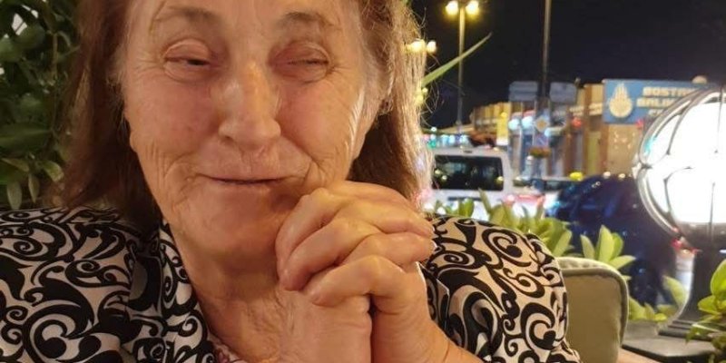 Cevriye Bayraktar, ellerini birleştirerek çenesinin altında tutuyor ve hafifçe gülümsüyor. Üzerinde siyah beyaz desenli bir ceket var. Arkasında yeşil bitkiler de bulunuyor.