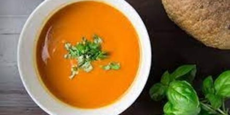 Ahşap bir masanın kenarında yeşil yapraklı bitki var. Masanın üzerinde beyaz çorba kasesinin içinde tarhana çorbası ve üzerinde doğranmış maydanoz var. Çorba kasesinin yanında yuvarlak tam buğday ekmeğinin yarısı görünüyor.