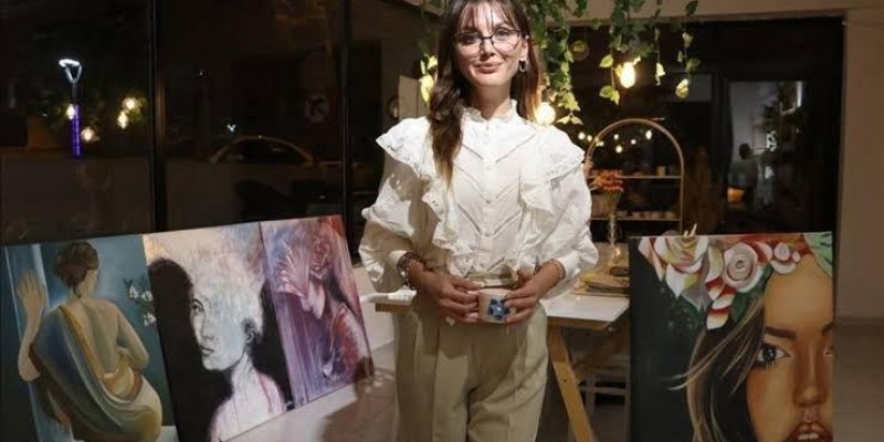 Bir sanat galerisinde veya sergi alanında duran genç bir kadın. Beyaz, fırfırlı bir bluz ve açık renk pantolon giymiş. Elinde bir fincan tutuyor ve gülümsüyor. Arkasında, üzerinde çeşitli resimler bulunan bir masa var. Resimlerden biri, sırtı dönük bir kadını ve başka bir resim, çiçeklerle süslenmiş bir kadının yüzünü gösteriyor. Ortamda loş bir aydınlatma var ve tavandan sarkan bitkiler dekorasyonu tamamlıyor. Arka planda cam bir duvar ve dışarıdaki gece manzarası.