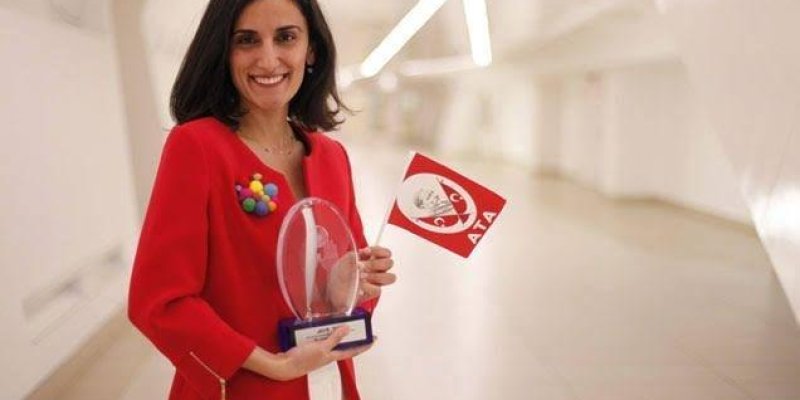 Canan Dağdeviren beyaz ve modern bir iç mekânda duruyor. Üzerinde kırmızı bir ceket ve beyaz bir pantolon var. Ceketin sol tarafında renkli bir broş bulunuyor. Saçları omuz hizasında, gülümsüyor. Elinde şeffaf bir ödül ve kırmızı-beyaz bir bayrak tutuyor. Bayrağın üzerinde Atatürk'ün resmi ve "ATA" yazısı yer alıyor.