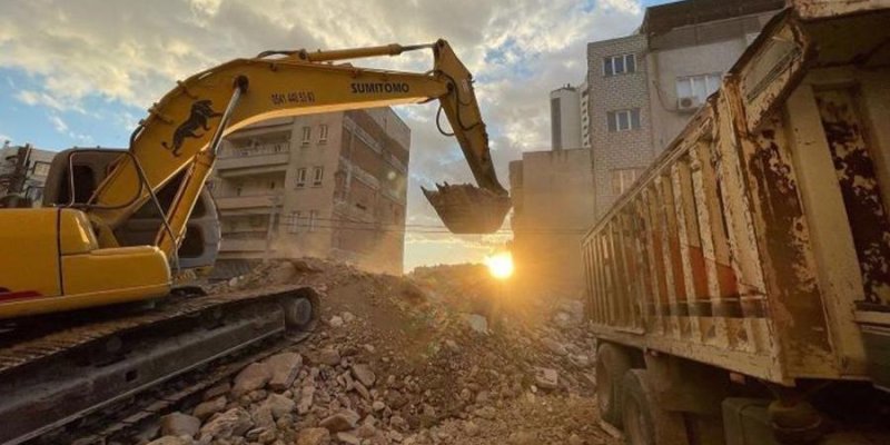 Güneş ışığında çalışan büyük bir sarı ekskavatör, moloz ve taş yığınlarının üzerinde duruyor ve kepçesi yukarıda, bir şey kaldırıyormuş gibi havada tutulmuş. Üzerinde “SUMITOMO” yazısı bulunan uzun kolu. Sağ tarafta, kasası paslı ve kirli görünen büyük bir kamyonun arka ve yan kısmı yer alıyor; kasanın içi görünmüyor. Ortada yerde, düzensiz yığılmış beton parçaları, taşlar ve inşaat molozları. Arka planda, çok katlı apartman binaları sıkışık şekilde yan yana duruyor; bazı pencerelerde klima ünit