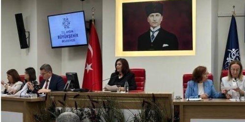 Bir belediye meclis/toplantı salonunda, uzun bir masanın arkasında oturan birkaç kişi ve duvarda büyük bir Atatürk portresi. Masanın ortasında siyah kıyafetli bir kadın mikrofon önünde konuşuyor gibi görünüyor; sağında ve solunda oturan kişiler kâğıtlara bakıyor veya not alıyor. Sol duvarda bir ekranda “BÜYÜKŞEHİR BELEDİYESİ” yazısı ve bir amblem görünüyor. Masanın arkasında Türk bayrağı ve sağ tarafta koyu renkli bir bayrak asılı. Ön planda izleyicilerin başları ve masanın önünde dekoratif bitk