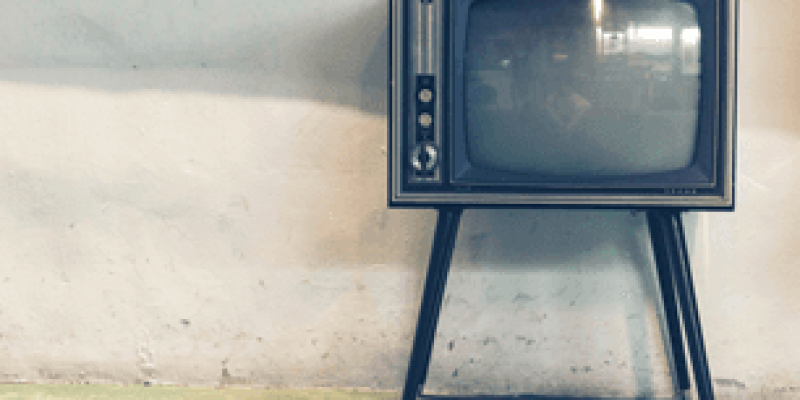 Açık bir mekanda beyaz badanalı duvara yaslanmış 4 ayaklı sehpa üzerinde eski siyah beyaz kapalı bir televizyon. Ekrana çevrenin görüntüsü yansımış. Televizyonun sol tarafında düğmeler var.