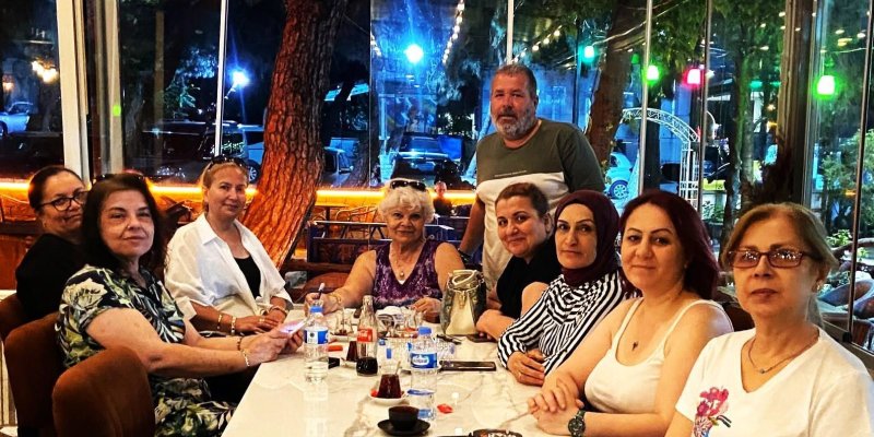 Restoran masasında oturan kalabalık bir arkadaş grubu. Uzun, parlak beyaz bir masanın etrafında dokuz kişi oturuyor, bir kişi ayakta duruyor; toplamda on yetişkin. Masada çay bardakları, su şişeleri, bir küçük cam Coca‑Cola şişesi, telefonlar, bir sürahi ve küçük tabaklar duruyor.