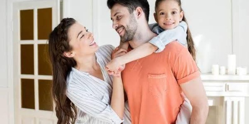 Mutlu bir aile. Ortada baba, sağında anne ve babanın sırtında bir kız çocuğu var. Baba turuncu bir tişört giymiş ve sakallı. Anne beyaz çizgili bir gömlek giymiş ve uzun saçlı. Kız çocuğu ise beyaz bir üst giymiş ve babasının sırtına sarılmış. Hepsi birbirine gülümseyerek bakıyor. Arka planda beyaz bir kapı ve beyaz bir duvar var.