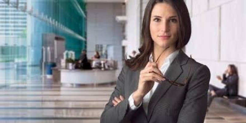 Modern bir ofis veya iş merkezi mekânında, ön planda bir kadın, koyu renkli bir takım elbise giymiş ve kollarını çaprazlamış şekilde duruyor. Elinde gözlük tutuyor ve doğrudan kameraya bakıyor. Arka planda, cam duvarlar ve büyük bir resepsiyon alanı görülüyor. Resepsiyonun arkasında birkaç kişi çalışıyor. Sağ tarafta duvara yaslanmış oturan iki kişi var.