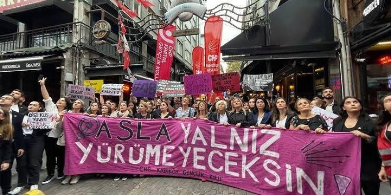 Kalabalık bir grup insan bir sokakta yürüyüş yapıyor. ellerinde çeşitli pankartlar taşıyorlar. En önde, büyük pembe bir pankart üzerinde "ASLA YALNIZ YÜRÜMEYECEKSİN!" yazıyor ve altında "CHP KADIKÖY GENÇLİK KOLU" ifadesi yer alıyor. Arkada, CHP (Cumhuriyet Halk Partisi) bayrakları ve flamaları görülüyor. Katılımcılar, genellikle kadınlardan oluşuyor ve çeşitli sloganlar içeren pankartlar taşıyorlar.