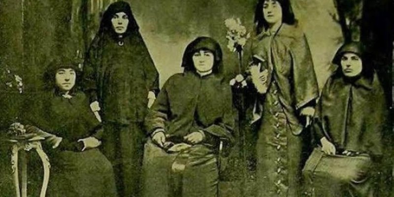Eski zamanlardan kalma bir grup kadın. Beş kadın, koyu renkli ve uzun elbiseler giymiş. Hepsi başlarını örtmüş ve ciddi bir ifadeyle kameraya bakıyorlar. Ortam, eski bir fotoğraf stüdyosunu andırıyor ve arka planda belirsiz bir dekor bulunuyor. Kadınlardan biri elinde çiçek tutuyor.