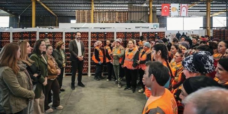 Depo benzeri geniş bir endüstriyel iç mekânda, iki tarafta karşılıklı duran kalabalık bir insan grubu. Ortada açık bir beton zemin koridoru; her iki yanda turuncu reflektörlü iş yelekleri giyen çok sayıda kişi sıraya yakın şekilde duruyor. Sol tarafta sivil giyimli birkaç kişi (ceketli ve montlu) gruba dönük duruyor. Orta kısımda takım elbiseli bir erkek, iki grubun arasında boşlukta ayakta duruyor. Arka planda beyaz raf sistemleri içinde kırmızı kasalarda/çekmecelerde istiflenmiş ürünler bulunu