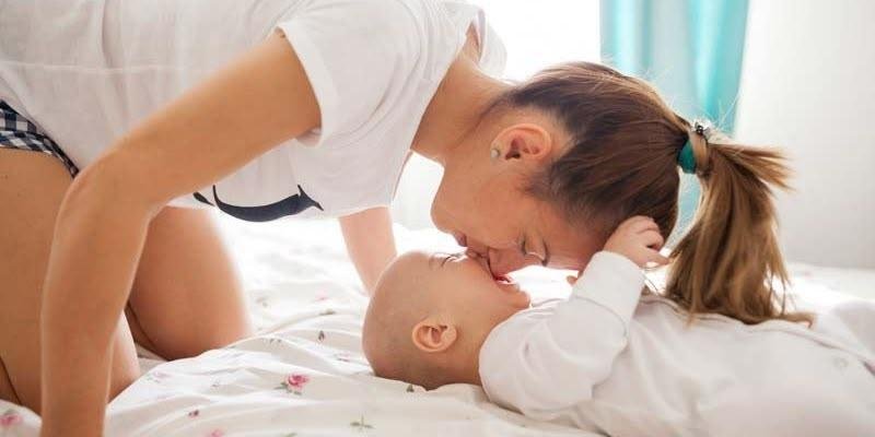 Yatakta yatan bir bebekle yüz yüze eğilmiş genç bir kadın beyaz bir tişört giymiş, açık tenli ve saçları arkadan toplanmış at kuyruğu şeklinde. Bebek açık tenli, beyaz bir tulum giymiş, sırtüstü yatıyor. Kadın, alnını bebeğin alnına değdiriyor ve yüzü çok yakın; aralarında sevgi dolu, şefkatli bir temas Bebeğin bir kolu yukarı kalkmış ve kadının yüzüne dokunur pozisyonda. Altlarında açık renk, çiçek desenli bir yatak örtüsü. Arka planda ışık alan bir pencere ve yanında mavi-turkuaz renkte bir pe