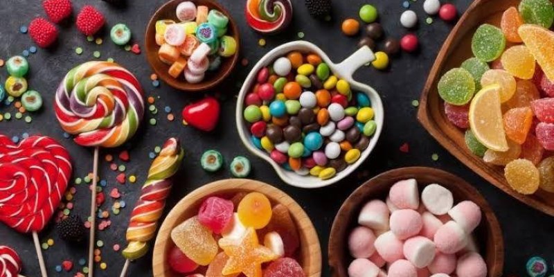 Çeşitli renkli şekerlemeler ve tatlılar. Görüntünün merkezinde, kalp şeklinde bir kâse içinde renkli drajeler bulunuyor. Etrafında farklı boyutlarda ve şekillerde kâseler var; bunların içinde jelibonlar, marshmallowlar ve meyve dilimi şeklinde şekerlemeler yer alıyor. Fotoğrafın sol tarafında renkli lolipoplar ve çubuk şekerler dikkat çekiyor. Arka planda küçük renkli şeker parçaları serpiştirilmiş.