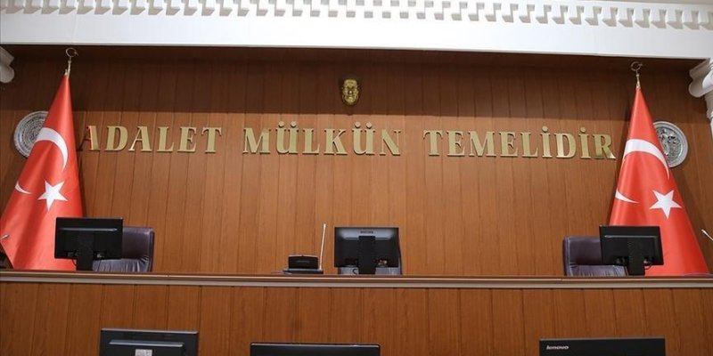 Ahşap kaplı bir mahkeme salonu kürsüsü. Arka duvarda büyük altın renkli harflerle “ADALET MÜLKÜN TEMELİDİR” yazısı görülüyor. Yazının üst orta kısmında küçük bir arma yer alıyor. Kürsünün solunda ve sağında birer Türk bayrağı asılı. Ön tarafta bilgisayar monitörleri ve hâkim koltukları görünüyor. Önde açık renk ahşap masalar ve sandalyeler var; masanın üzerinde siyah bir telefon duruyor. Arkada raflarda çok sayıda dosya/klasör ve kitap dizili. Sağ duvarda büyük bir Atatürk portresi asılı. Tavand