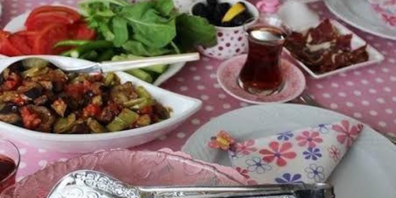 Pembe puantiyeli bir masa örtüsü ile kaplanmış bir kahvaltı masasında çeşitli yiyecekler ve içecekler bulunuyor. Önde bir tabakta kızarmış patates var ve üzerinde çatal ve bıçak yer alıyor. Masanın diğer kısımlarında domates, salatalık, yeşillikler, siyah ve yeşil zeytinler, peynir çeşitleri, kuru meyveler ve ceviz gibi kahvaltılık ürünler, birkaç çay bardağı bulunuyor.