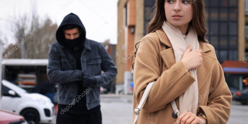 Şehir dışında çekilmiş bir sokak sahnesinde, ön planda açık tenli bir kadın ve arka planda kapüşonlu bir erkek. Kadın sağ tarafta; uzun kahverengi saçlı, bej bir palto ve açık renk bir atkı takıyor. Omzunda açık renk bir çanta askısı ve bir eli çantanın üstünde, diğer eli göğsüne yakın şekilde paltonun yakasını/atkısını tutuyor; başını sağa çevirip endişeli, tedirgin bir ifadeyle yana bakıyor. Arka planda sol tarafta bir erkek; siyah kapüşonlu üst giymiş, yüzünün alt kısmını siyah bir kumaşla ka