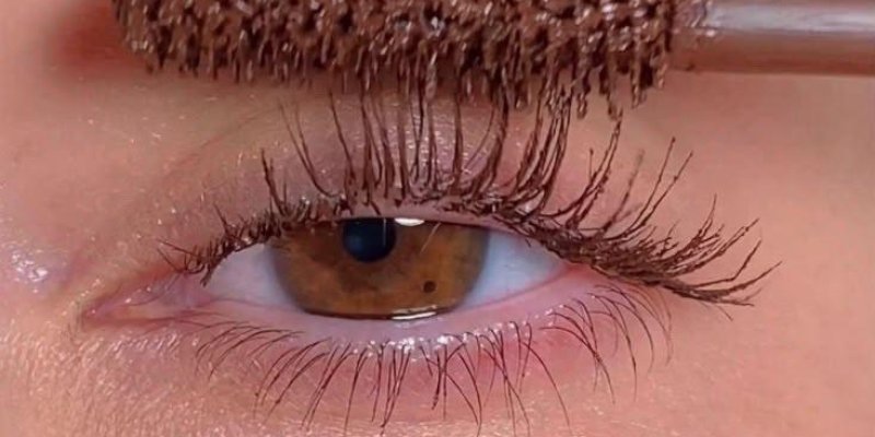 Kahverengi irise sahip bir göz ve üst kirpiklerin üzerinde duran bir maskara fırçası. Göz kapakları ve çevresindeki cilt parlak ve nemli duruyor, aydınlatıcı ya da nemlendirici uygulanmış. Maskara fırçasındaki ürün açık-koyu arası bir kahverengi tonunda ve fırça kılları yoğun, kalın. Üst kirpikler uzun, tek tek ayrılmış ve maskara ile belirginleşmiş; uçlara doğru hafif yukarı doğru kıvrım. Alt kirpiklerde de hafif maskara etkisi, ancak üst kirpiklere göre daha hafif. Kaşlar doğal, seyrek olmayan