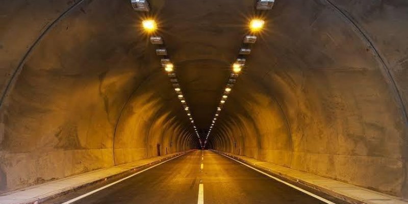 Aydınlatılmış bir karayolu tüneli. Tünelin içinden düz bir şekilde bakılıyor. Tünelin tavanında sıralı şekilde yerleştirilmiş sarı ışıklar var ve bu ışıklar tünelin içini aydınlatıyor. Yolun ortasında beyaz bir şerit bulunuyor. Yolun iki yanında kaldırım benzeri dar alanlar mevcut. Tünelin duvarları beton ve hafifçe sarımsı bir ışıkla aydınlanmış. Tünelin sonunda uzak bir noktada karanlık görünüyor, çıkış veya diğer ucu net şekilde görünmüyor. Yolda hiç araç veya insan yok, tamamen boş bir tünel