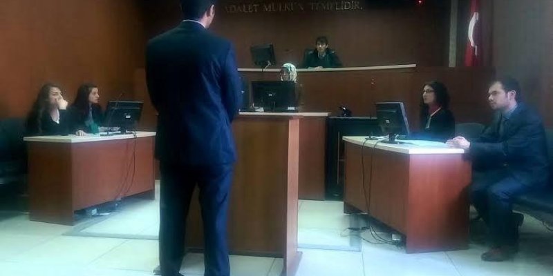 Bir mahkeme salonu. Ortada bir avukat veya savcı ayakta duruyor ve kürsüye doğru bakıyor. Kürsünün arkasında bir hâkim, sağ tarafta iki kişi oturuyor, biri bilgisayar ekranına bakıyor. Sol tarafta iki kişi daha oturuyor, onların da önünde bir bilgisayar var. Duvarın arkasında "Adalet mülkün temelidir." yazısı ve bir Türk bayrağı görülüyor.