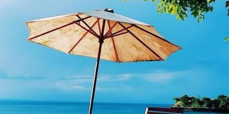 Kumsalda bir şezlong ve büyük bir şemsiye açık ve şezlongun yanında duruyor. Kumlar beyaz ve deniz oldukça sakin. Gökyüzü açık mavi ve birkaç bulut var. Üst kısımda yeşil yapraklı ağaç dalları görünüyor. Arka planda denizin üzerinde küçük bir ada veya kara parçası var.