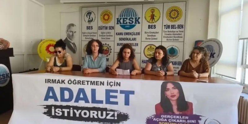 Masa arkasında oturan beş kadın. Ortadaki bir kâğıt tutuyor ve okuyor, diğerleri yan yana oturmuş, önlerinde büyük bir pankart. Kadınların hepsi yetişkin, saçları omuz hizasında ya da daha uzun; soldan ilki açık tenli, siyah üst ve güneş gözlüğü takmış, saçları tepede toplanmış. Yanındaki açık tenli, dalgalı kahverengi saçlı, açık mavi bir üst giymiş. Ortadaki açık tenli, dalgalı koyu kahverengi saçlı, kolsuz gri bir üst giymiş. Dördüncü açık tenli, dalgalı koyu kahverengi saçlı, desenli kısa ko