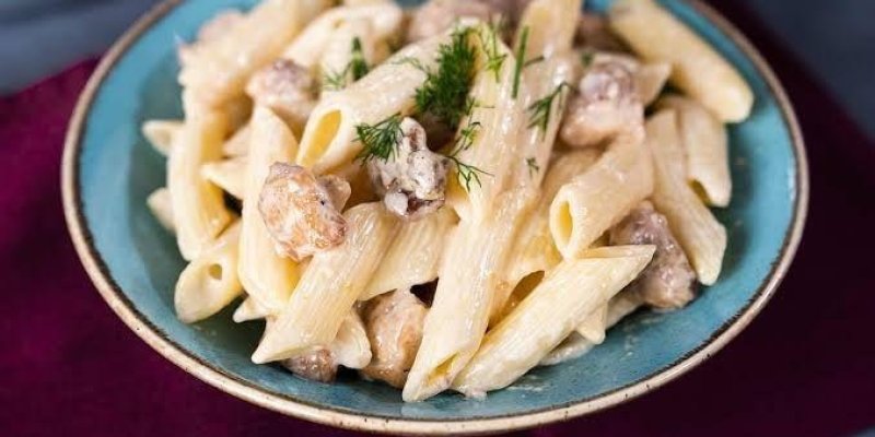 Mavi bir tabakta sunulmuş kremalı bir makarna yemeği. Makarna penne şeklinde ve kremalı bir sosla kaplanmış. İçerisinde küçük küpler halinde kesilmiş tavuk parçaları var. Üzerine taze dereotu serpiştirilmiş. Tabak, bordo bir örtünün üzerinde duruyor.