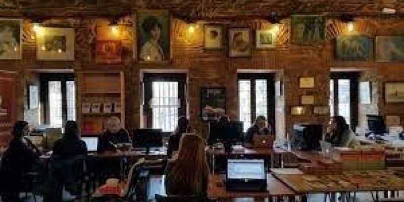 Kütüphane cafe ortamında oturan kadınlar ve erkekler var. Birçok masa, kitap, duvarlar ışıklarla aydınlatılmış, duvarlarda resimler var.