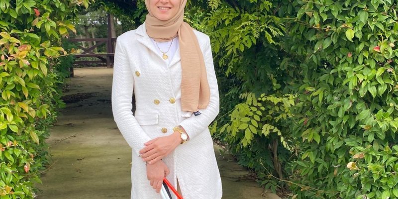 Esra Durmuş, yeşil yapraklarla kaplı bir çalı kemerinin altında duruyor. Açık kahverengi başörtüsü takıyor ve beyaz, düğmeli bir kaban/ceket ile açık renk pantolon giymiş; ayağında beyaz spor ayakkabılar var. Bileğinde saat ve boynunda küçük bir kolye görünüyor. Ellerini önde birleştirmiş ve kırmızı-beyaz renkli katlanır bir baston tutuyor. Zemin beton bir yürüyüş yolu; arka planda bahçe içinde bir geçit ve uzakta ahşap bir yapı seçiliyor. Gökyüzü gri bulutlu.