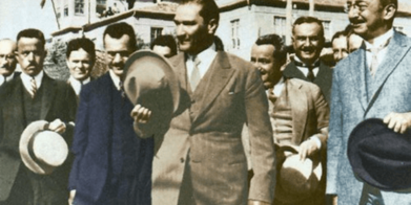 Toprak rengi takım elbiseli, beyaz gömleği ve elinde toprak rengi şapkası ile Atatürk ve yanında mavi, lacivert, siyah takım elbise giyinmiş birkaç erkek ve arkada sadece kafası görünen kısa saçlı bir kadın duruyor.