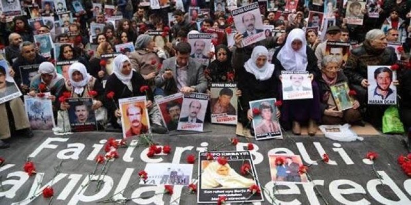 Bir grup insan bir protesto veya anma etkinliğinde toplanmış. Çoğunluğu kadın olan bu insanlar, ellerinde kayıp veya öldürülmüş yakınlarının fotoğraflarını tutuyorlar. Bazı kadınlar başörtüsü takıyor. Yerde büyük bir pankart var ve üzerinde "Failleri belli kayıplar nerede?" yazıyor. Pankartın etrafında kırmızı karanfiller yer alıyor. Kayıp yakınlarını anmak ve adalet talep etmek amacıyla düzenlenen Cumartesi Anneleri eylemi.
