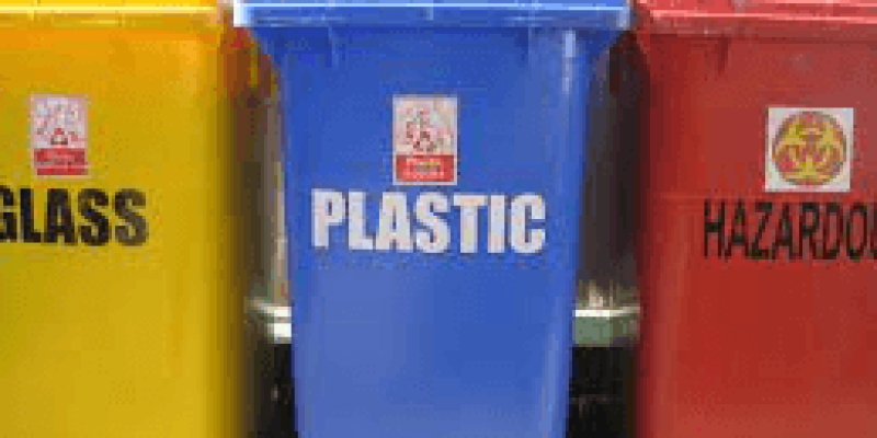 Üç tane üyük boy plastik çöp kutusu yan yana duruyor. En soldaki sarı kutunun gövdesinde siyah büyük puntolarla İngilizce “glass”, “cam” yazıyor. Yanındaki mavi kutunun gövdesinde büyük puntolarla İngilizce “Plastic”, “Plastik” yazıyor. Sondaki kırmızı kutunun gövdesinde“Hazardous”, “Tehlikeli” yazıyor.