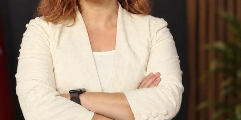 Açık tenli, kızıl dalgalı saçlı, kolları göğsünde çapraz duruyor. Yüzünde hafif, kendinden emin bir gülümseme ve doğrudan kameraya dönük. Üzerinde krem rengi, dokulu bir blazer ceket, içinde beyaz bir tişört ve mavi kot pantolon. Ceket kollarının uçlarında üçer düğme detayları. Sol bileğinde koyu renk bir akıllı saat ya da bileklik takılı. Kulaklarında açık renk sallantılı küpeler.