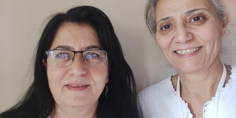 İki kadın. Soldaki kadın siyah saçlı ve gözlük takıyor. Yeşil bir üst giymiş. Sağdaki kadın ise beyaz saçlı ve beyaz bir bluz giymiş. İkisi de kameraya bakarak gülümsüyorlar. Arka plan sade ve bej renginde.