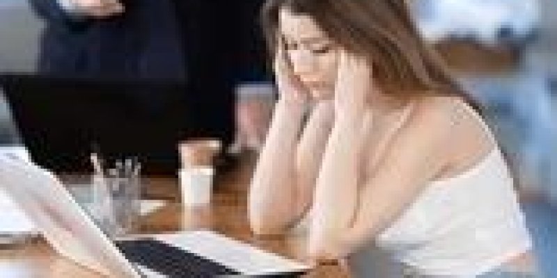 Açık ofis ortamında ahşap bir masada oturan genç kadın beyaz askılı bir bluz ve açık mavi kot pantolon giymiş, dirseklerini masaya dayamış, ellerini iki yana başına götürmüş ve başını hafifçe öne eğmiş. Önünde açık bir dizüstü bilgisayar, yanında birkaç kâğıt ve bir kalem. Yüz ifadesi yorgun veya sıkılmış gibi, omuzları düşük duruyor. Arka planda koyu renk takım elbise giymiş, gri saçlı bir erkek ayakta duruyor; bir eli açık şekilde öne doğru uzanmış, sanki konuşuyor veya bir şey anlatıyor gibi