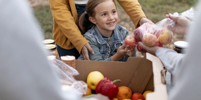Gıda yardımı sırasında meyve-sebze dolu bir karton kutunun önünde duran, gülümseyen küçük bir kız çocuğu. Açık tenli; kahverengi saçları iki yandan toplanmış, üzerinde kot yelek ve uzun kollu bir üst bulunuyor. Arkasında hardal sarısı hırka giymiş bir yetişkin duruyor ve kızın omuzlarına/yanına destek olur şekilde elleri görünüyor. Sağ tarafta bir başka kişi, kızın uzattığı poşete doğru içinde kırmızı ve sarı elmalar olan şeffaf bir poşet veriyor. Ön plandaki kutunun içinde kırmızı dolmalık bibe