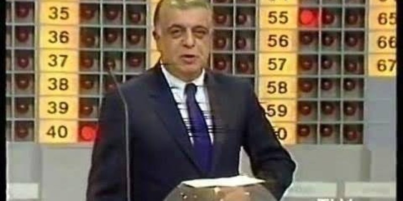 Bir televizyon stüdyosunda çekilmiş sahne. Ortada bir adam, takım elbise giymiş ve kravat takmış bir şekilde duruyor. Önünde, şeffaf bir küre içinde bir nesne var. Arka planda, üzerinde 1'den 60'a kadar sıralanmış sayılar bulunan bir pano. Panonun bazı bölümleri kırmızı renkle işaretlenmiş. Ekranın sağ alt köşesinde "TV1" logosu.