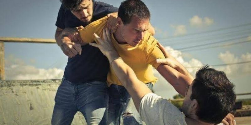 Üç erkek, ortadaki sarı bir tişört giymiş ve öne doğru eğilmiş. Sağdaki yere düşmüş ve beyaz bir tişört giymiş. Sol taraftaki koyu renk bir tişört giymiş ve ortadaki adamı tutuyor. Ortadaki adam, sağdaki adama doğru uzanmış ve ona doğru hamle yapıyor.