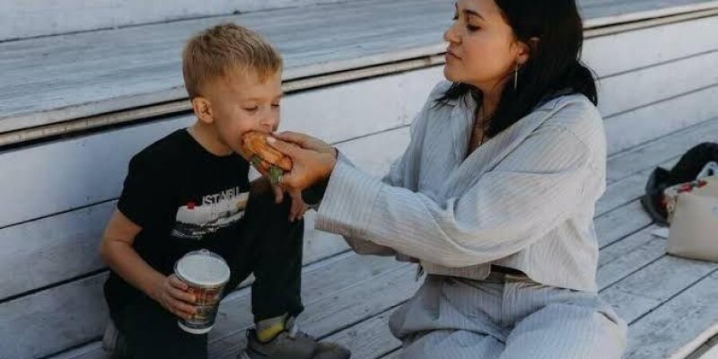 Basamaklı ahşap bir oturma alanında yan yana oturan bir kadın ve küçük bir erkek çocuk. Kadın hafif açık renkli, bol, pijama tarzı bir takım giymiş, koyu renk uzun saçlı ve beyaz tenli. Yan tarafında oturan çocuğun ağzına iki eliyle uzun bir sandviç tutarak yediriyor. Çocuk sarı saçlı, açık tenli, siyah tişört ve koyu pantolon giymiş, ayaklarında gri spor ayakkabılar. Sol elinde kapaklı, karton bir içecek bardağı tutuyor; içecek büyük boy görünüyor. İkisi de aynı yöne dönük, çocuk otururken bira