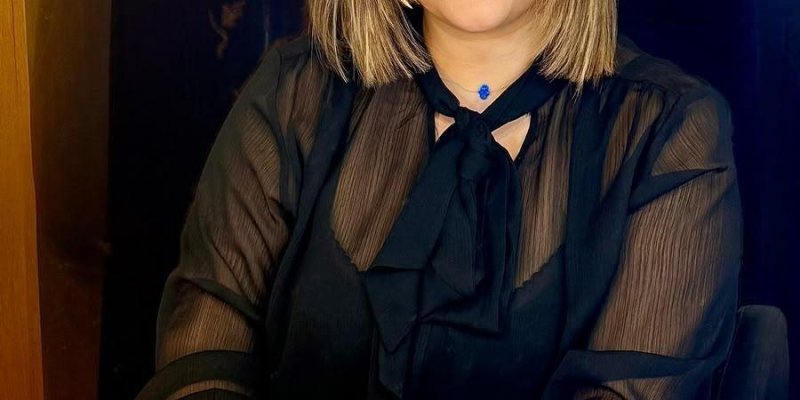 Ofis ortamında beyaz masanın başında oturan Avukat Süreyya Yarli. Ten rengi açık, yüzünde doğal ama belirgin bir makyaj; kaşları belirgin, gözlerinde hafif eyeliner ve far, dudaklarında mat bir ruj. Saçları omuz hizasında, düz, açık kahverengi tonunda ve açık renk ışıltılarla boyanmış, ortadan ayrılmış. Üzerinde transparan kollu, yakasında büyük fiyonk detayı olan siyah bluzun içinden siyah bir iç parça görünüyor. Boynunda küçük, mavi taşlı zarif bir kolye. Elleri masanın üzerinde birbirinin üze