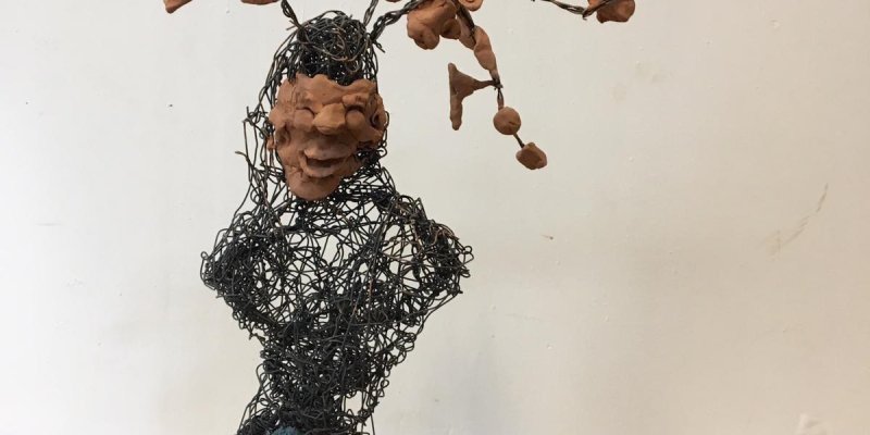 Bir sanat eseri, metal telden yapılmış bir iskelet üzerine yerleştirilmiş kil parçalarından oluşuyor. Üst kısımda, tel iskeletin üzerinde bir yüz şekli var ve bu yüzün üst kısmından dallar gibi uzanan kil parçaları çıkıyor. Alt kısımda, mavi ve turuncu renklerde kil parçaları, eserin tabanını oluşturuyor. Eser, ahşap bir yüzey üzerinde sergileniyor.